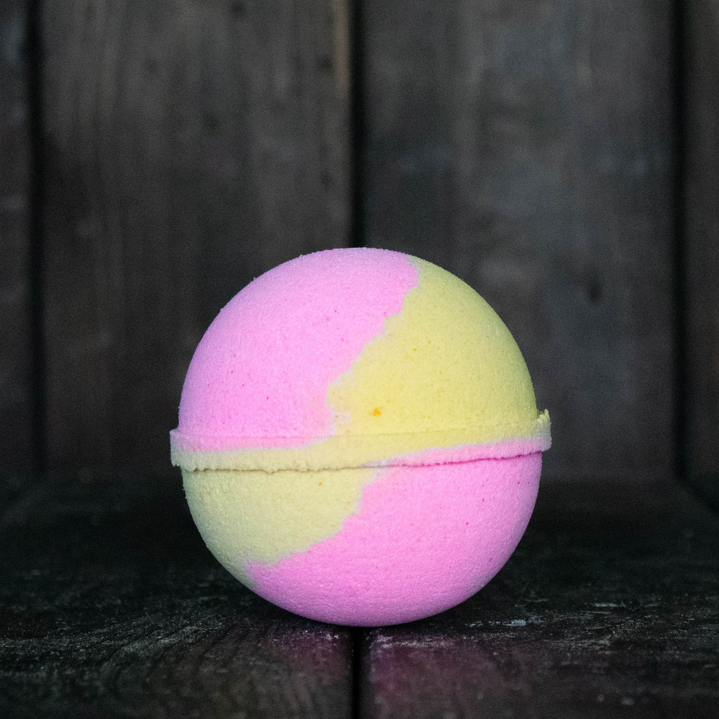 A pink and yellow bath bomb in front of a dark wood background.