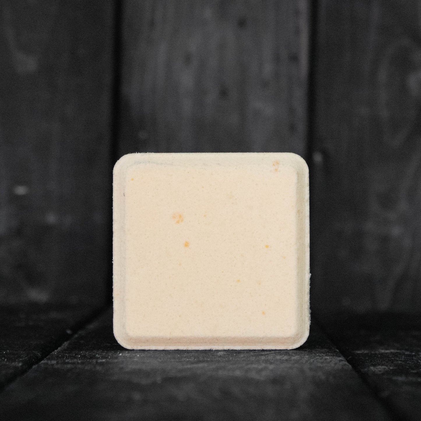 A light orange, square shaped shower steamer in front of a dark wood background.