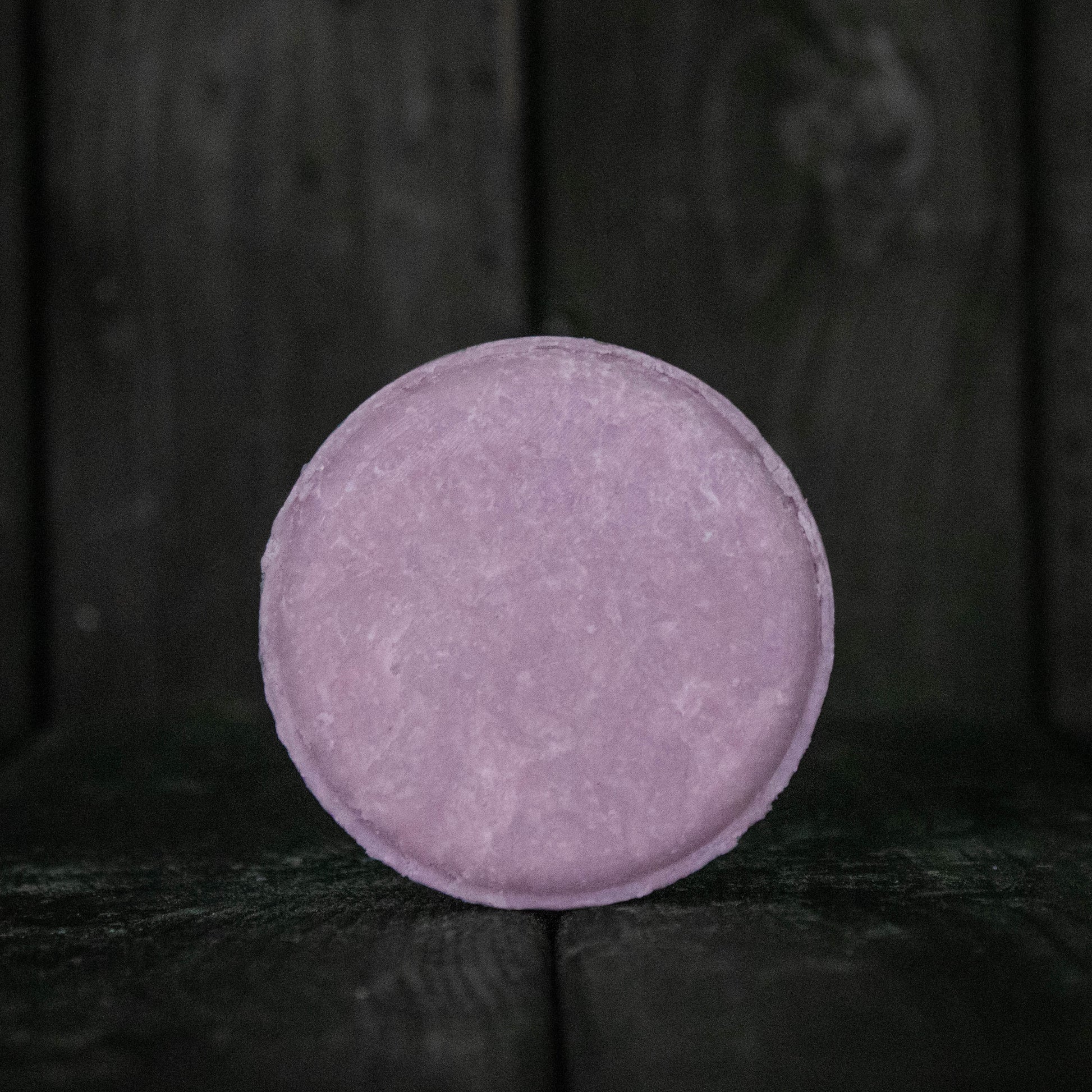 A round purple shampoo bar in front of a dark wood background.