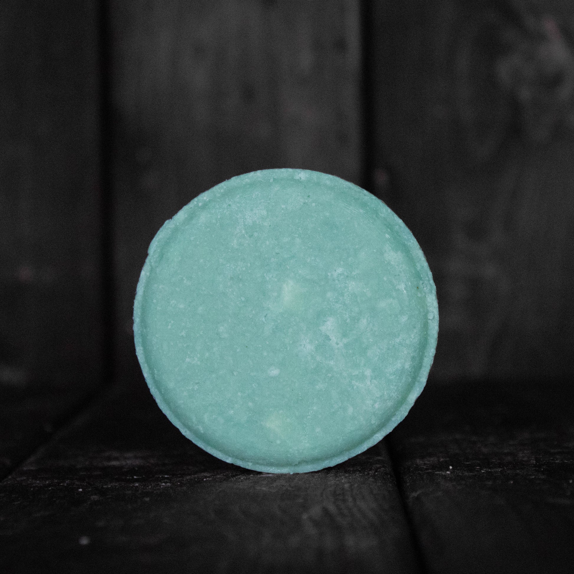 A round turquoise shampoo bar in front of a dark wood background.